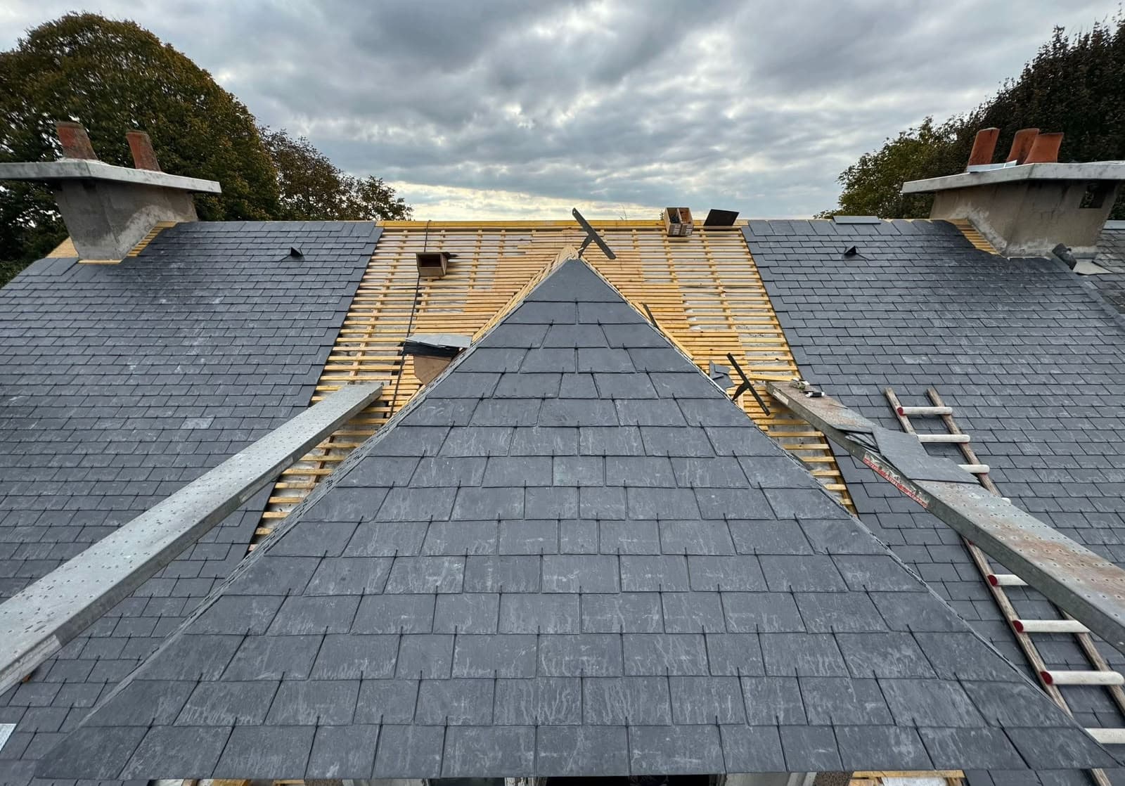 Pose de tuiles d'ardoise grise sur une toiture en rénovation sous un ciel couvert.