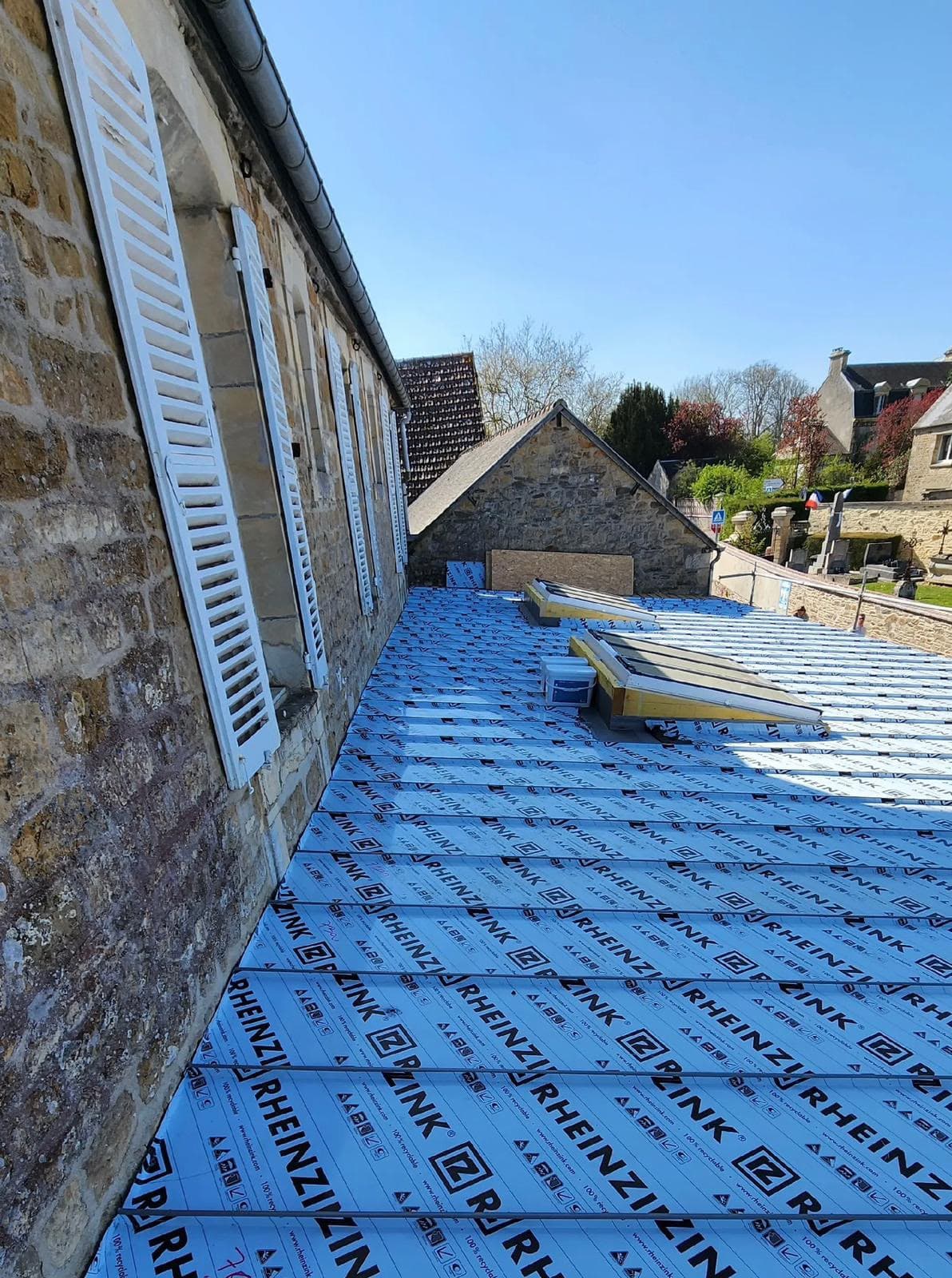 Membrane de toiture Rheinzink bleue sur un bâtiment en pierre avec des volets blancs.